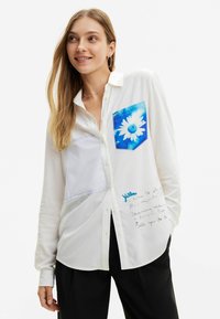 Camicia bianca con bottoni, caratterizzata da una tasca floreale blu e un pannello striato in alto a sinistra, con dettagli di testo scritto a mano in basso a destra.