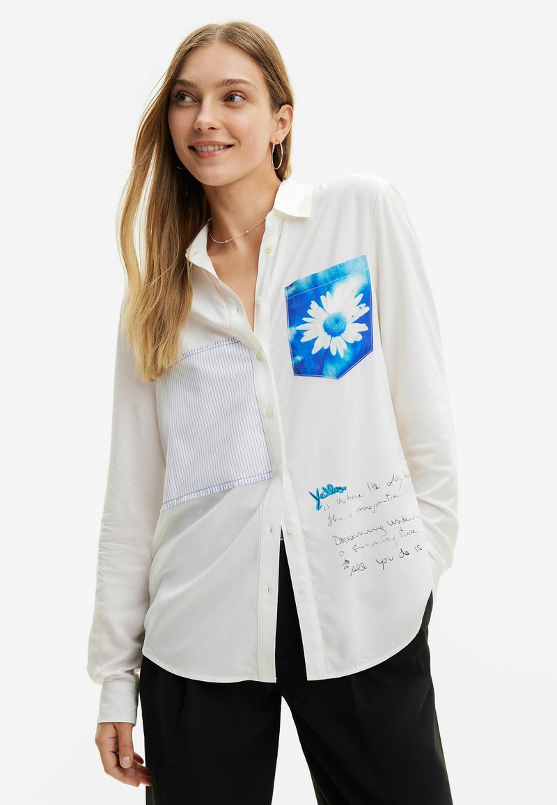 Camicia bianca con bottoni, caratterizzata da una tasca floreale blu e un pannello striato in alto a sinistra, con dettagli di testo scritto a mano in basso a destra.
