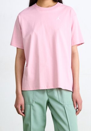 Persoon die een pastelroze t-shirt met korte mouwen draagt, voorzien van een klein wit logo op de borst, en losse lichtgroene broek.