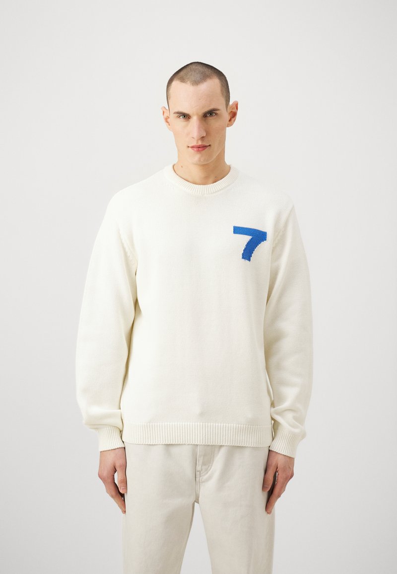 Pull en tricot blanc avec un design bleu "7" sur la poitrine gauche, doté d'un col rond, de poignets et d'un ourlet côtelés.