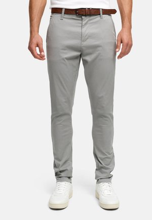 Man draagt lichtgrijze slim-fit broek met een bruine leren riem en witte sneakers, staand tegen een effen achtergrond.