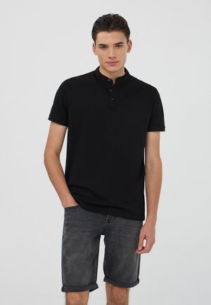 Polo nero con colletto alto, dotato di patta con bottoni, abbinato a pantaloni corti in denim grigi arrotolati all'orlo.