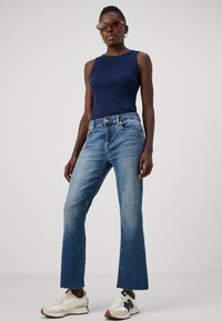 Marinblå utanärmad topp, ljusblå denimbyxor med utsvängd fot och fransad nederkant, samt vita sneakers med bruna detaljer. Transparenta solglasögon bärs.