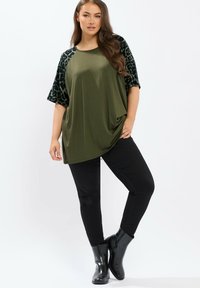 Vrouw in een olijfgroene oversized top met zwarte luipaardprintmouwen, zwarte skinny broek en zwarte enkellaarsjes, poseert tegen een witte achtergrond.