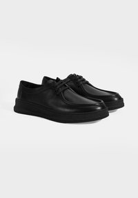 Schwarze Lederschuhe mit abgerundeter Spitze, Schnürdesign und einer dicken schwarzen Gummisohle. Glatte Textur mit minimalen nahtdetails.