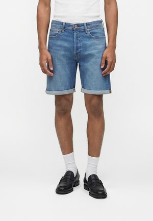 Bărbat purtând pantaloni scurți albastre din denim suflecați, șosete albe, mocasini negri din piele și o cămașă albă, stând într-un fundal simplu.