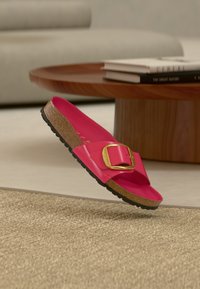 Ροζ σανδάλι με γυαλιστερό φινίρισμα, με μεγάλο χρυσό πόρπη, πάτο από φελλό και υφασμένη μαύρη σόλα, αιωρούμενο  πάνω από ένα καφέ χαλί.