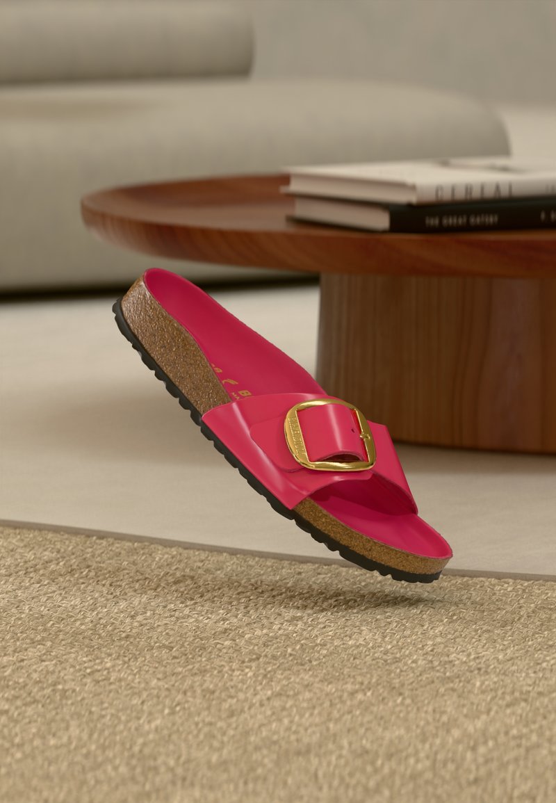 Lyserød slidesandal med blank finish, der har en stor guldbeslag, kork-fodbed og tekstureret sort sål, hængende over et beige tæppe.