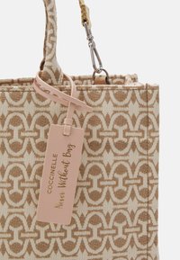 Torebka Coccinelle w beżowo-brązowym tkanym materiale, z powtarzającym się wzorem oraz różową metką z napisem "Nigdy bez torby" w złotym kolory.