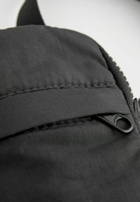 Pochette en tissu noir avec zip horizontal et un petit onglet en tissu. Texture lisse avec des coutures visibles le long des bords.