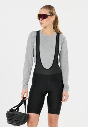 Frau in grauem Langarmshirt und schwarzer Radhose mit Trägern, die einen schwarzen Helm hält und große Sportsonnenbrillen trägt.