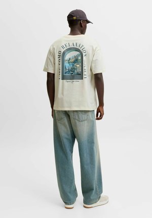 Ung mand iført en cremefarvet t-shirt med en farverig søscene og tekst på ryggen, løstsiddende blå jeans, hvide sneakers og en mørk kasket.