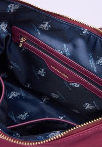 Interno della borsa bordeaux con rivestimento blu navy, dotato di una tasca con zip e un motivo con logo stampato. Maniglia della zip in tonalità oro e etichetta impressa.
