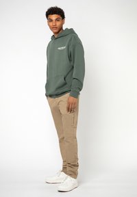 Grüne Baumwollkapuzenjacke mit Fronttasche und weißem Logo, kombiniert mit beigen Hosen und weißen Turnschuhen, präsentiert an einem stehenden Model.