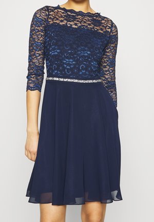 Nainen, jolla on polvipituinen tummansininen mekko, jossa on pitsiset kolmen neljänneksen hihat, läpikuultava pitsitoppi ja hopeinen vyö vyötäröllä.