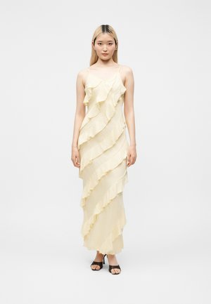 Νεαρή γυναίκα φορεί μακρύ, κρεμ φόρεμα με βολάν και λεπτές τιράντες, καθώς και μαύρα ανοιχτά τακούνια, στέκεται μπροστά σε απλό φόντο.