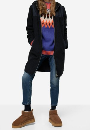 Personne portant un pull coloré à motifs, un long manteau noir zippé, un jean bleu, des chaussettes noires et des bottines marron en daim.