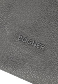 Sivkasta teksturirana usnje z utisnjenim logotipom "BOGNER". Ima kamenčkast površinski vzorec, kar dodaja taktilni kontrast celotni zasnovi.