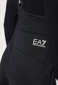 Juodi sportiniai džinsai, pagaminti iš lygios, tempiančios medžiagos, su išskirtiniu EA7 Emporio Armani logotipu ant nugaros. Siūlių detalės suteikia subtilaus dizaino.