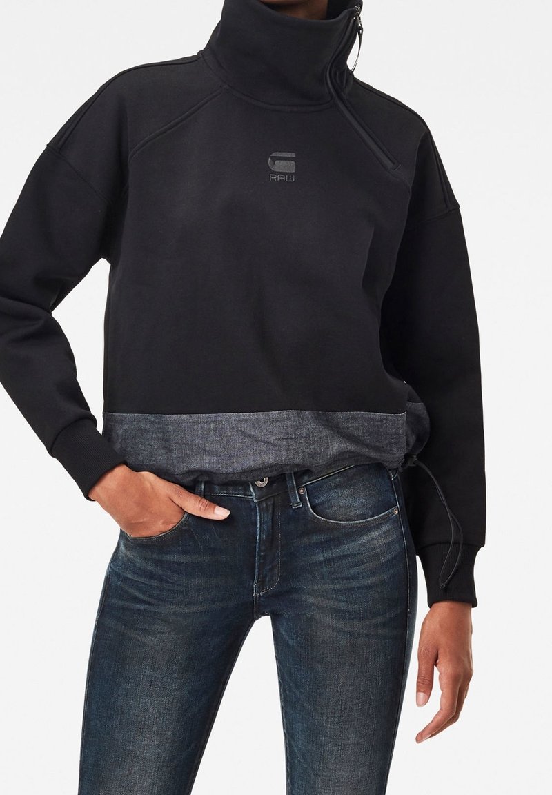 Personne portant un pull noir à col montant avec une fermeture éclair à l'avant et un logo, associé à un jean en denim bleu foncé, main dans la poche.