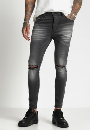 Mand iført slim-fit, falmede sorte revnede jeans med horisontale revner ved knæene, sorte sneakers, hvid skjorte og synlige arm tatoveringer.