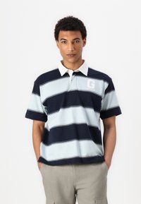 Polo majica s kratkimi rokavi z mornarsko modrimi in svetlo modrimi črtami. Ima bel ovratnik in logotip na prsih. Narejena je iz mehkega blaga.