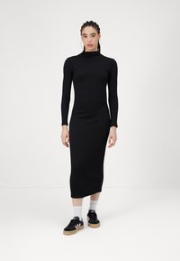 Μαύρο πλεκτό φόρεμα με ριμπ ύφανση, μακριά μανίκια, ψηλή λαιμόκοψη και εφαρμοστή γραμμή; συνδυασμένο με μαύρα αθλητικά παπούτσια και λευκές κάλτσες.