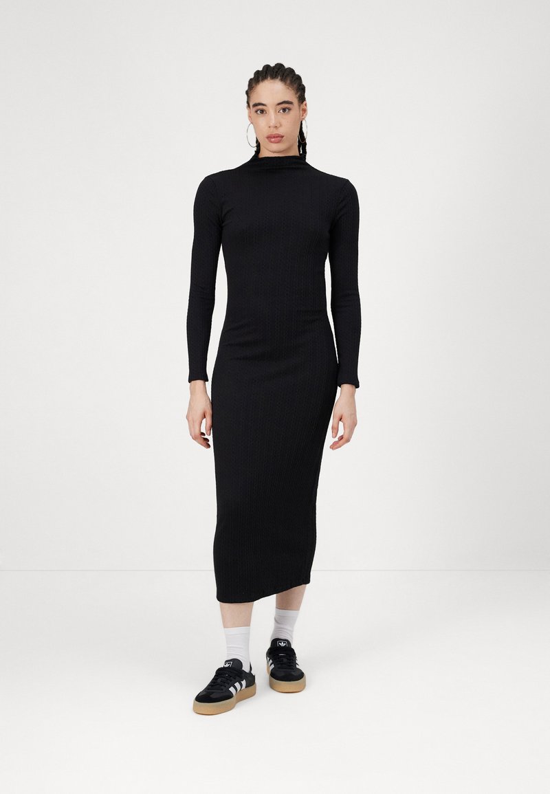 Μαύρο πλεκτό φόρεμα με ριμπ ύφανση, μακριά μανίκια, ψηλή λαιμόκοψη και εφαρμοστή γραμμή; συνδυασμένο με μαύρα αθλητικά παπούτσια και λευκές κάλτσες.