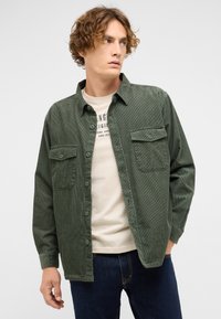 Grünes Cordhemd mit Knopfverschluss, zwei Brusttaschen und strukturierter Oberfläche, getragen über einem hellbeigen T-Shirt und Jeans.