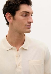 Camisa polo beige hecha de material suave, con un collar, tres botones y un bolsillo de parche en el lado izquierdo del pecho. Sin patrones ni acentos visibles.