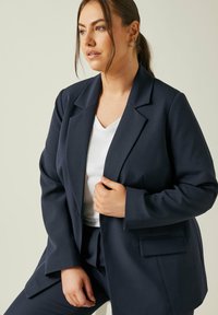 Granatowa marynarka o strukturalnym kroju, z wykładanymi klapami i jednym guzikiem. W połączeniu z białą koszulą w serek.