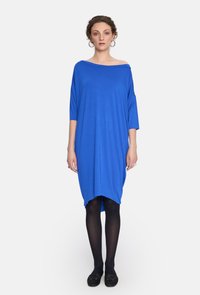 Luźna, nieco odkrywająca ramiona niebieska sukienka z miękkiego materiału, z rękawami do łokci i asymetrycznym dołem, zestawiona z czarnymi rajstopami i butami.