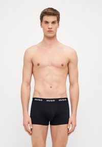 Fekete pamut boxer alsónadrág, amely szoros illeszkedésű és fekete derékkal rendelkezik, amelyen a „HUGO” logó fehér színben található. A modell semleges pózban áll.