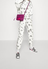 Person som bär grå och vit tie-dye sweatpants och sweatshirt, grå och vita sneakers med röda accenter, samt en liten lila crossbody-väska.