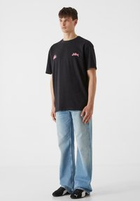 Must musta T-särk roosa graafiliste detailidega, koos helesiniste laienevate teksade ja mustade spordijalatsitega. Üldiselt lõdva riietuse disain.