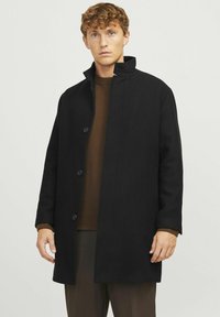 Abrigo de lana negra con un cuello alto, cierre de botones en la parte delantera y bolsillos laterales. Textura suave; diseño entallado, llevado sobre un suéter marrón.