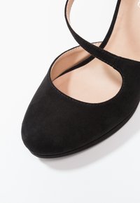 Chaussure en daim noir avec un bout arrondi, des découpes sur les côtés et une sangle à la cheville. Doublure intérieure lisse de couleur claire.