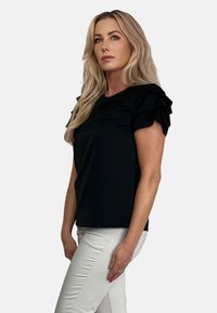 Czarna koszulka z krótkim rękawem z falbaniastymi akcentami na ramionach. Wykonana z gładkiego materiału, o luźnym kroju. Noszona z jasnymi spodniami.