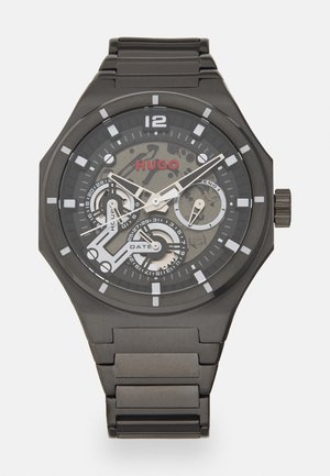 Black stainless steel watch with a round face, white hour markers, and a camouflage pattern. Features date and chronograph sub-dials. Red "HUGO" logo.