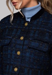 Marinblå tweedjacka med guldfärgade knappar, två framtill liggande fickor och en krage i sammet. Den texturerade tyget har ett subtilt rutig mönster.