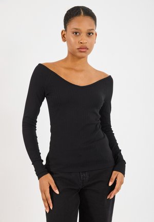 Langermet svart ribbestrikket topp med off-shoulder utringning, tettsittende silhuett og myk tekstur, parret med svarte bukser.