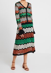 Kvinna som bär en knappknäppt långärmad cardigan och matchande midikjol med flerfärgade zigzag-mönster i grön, brun, svart och vit.