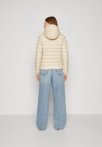 Beige polstret jakke med hette, med horisontalt quiltet design. Kombinert med vide lyseblå jeans, som skaper et uformelt utseende.