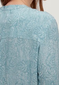 Frau, die ein hellblaues Kleid mit weißen abstrakten Blumen- und Strichmustern trägt, von hinten und im Schulterbereich gezeigt.