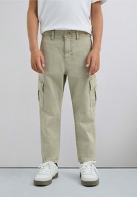 Pantalones cargo caqui con dos bolsillos laterales, hechos de una tela suave, corte recto y cierre de botón. Combinados con zapatillas blancas.