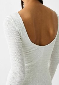 Robe blanche texturée, ajustée, avec manches longues et un profond décolleté dans le dos, dotée d'un design smocké qui ajoute de la profondeur et du détail.