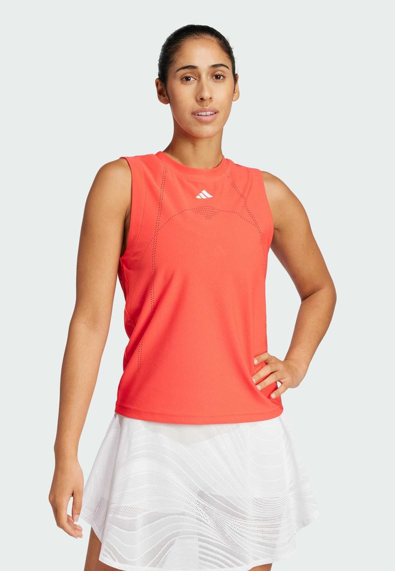 Rdeča atletska majica brez rokavov z okroglim izrezom, perforiranimi detajli in adidasovim logotipom. Kombinirana z belo krilom z valovitimi vzorci.