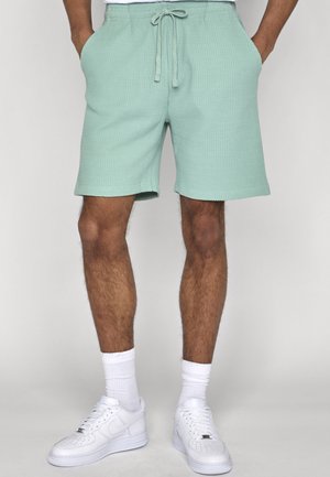 Muž oblečený v mätovo zelených textúrovaných šortkách so šnúrkou, bielych ponožkách a bielych teniskách Nike, stojaci s rukami vreckách.