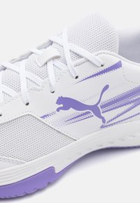 Hvit atletisk sko med mesh-overdel, lilla aksenter og en hoppende katt-logo. Har en teksturert såle og kontrasterende lilla hæl.