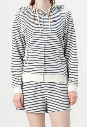 Personne portant un sweat à capuche blanc et un short à rayures horizontales noires, sweat à capuche fermé par une fermeture éclair, manches longues, et mains détendues le long du corps.